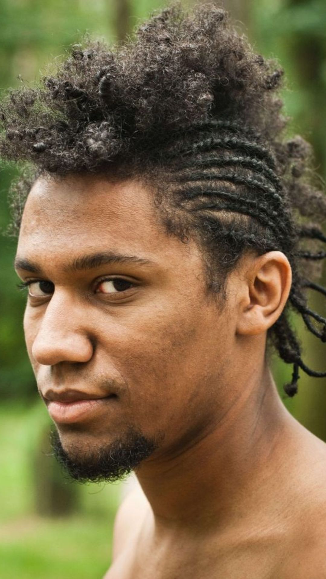 A young man with dreadlocks poses in the woods, highlighting his intricate braid hairstyle against a backdrop of trees.